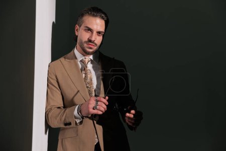 Photo for Attractive young businessman in brown suit taking sunglasses down and looking forward in front of grey background - Royalty Free Image