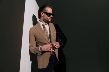 Photo for Confident elegant businessman with sunglasses looking to side and buttoning brown suit in front of grey background - Royalty Free Image