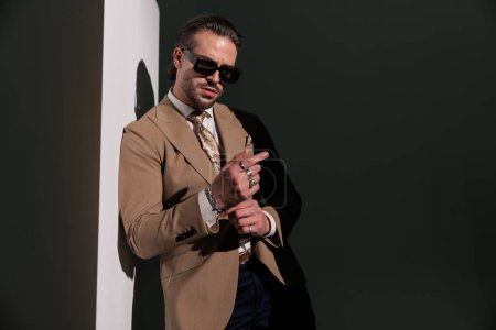 Foto de Retrato de un joven hombre de negocios elegante con gafas de sol ajustando pulseras, mirando hacia adelante y acostado en una pared gris - Imagen libre de derechos