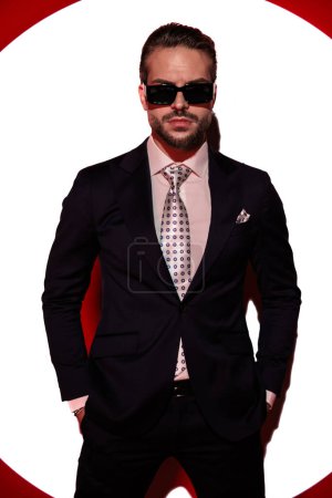 Téléchargez les photos : Portrait de jeune homme confiant avec des lunettes de soleil tenant la main dans les poches et regardant vers l'avant tout en posant devant les projecteurs - en image libre de droit