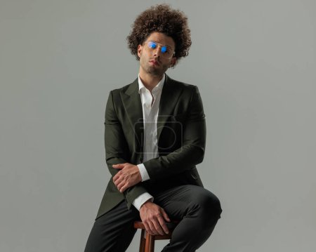 Téléchargez les photos : Sexy jeune homme d'affaires en costume vert avec des lunettes assis sur une chaise en bois, touchant les bras et regardant vers l'avant devant fond vert - en image libre de droit