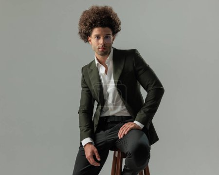 Téléchargez les photos : Homme d'affaires attrayant en costume vert assis avec la paume sur la cuisse, regardant vers l'avant en face de fond vert foncé - en image libre de droit