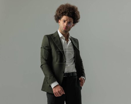 Téléchargez les photos : Jeune homme d'affaires sérieux en costume vert avec chemise à col ouvert et lunettes tenant la main dans la poche devant fond gris - en image libre de droit