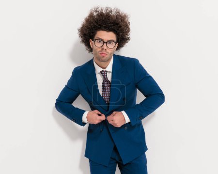 Photo for Portrait of handsome young man with glasses and curly hair buttoning navy blue suit in front of grey background - Royalty Free Image