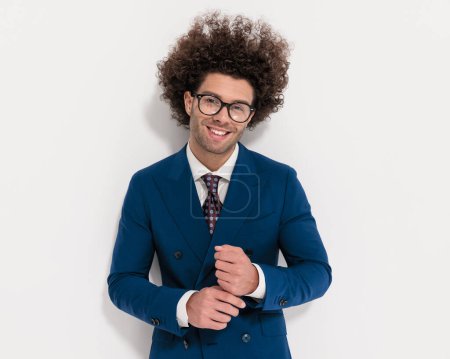 Foto de Feliz joven hombre de negocios con gafas en traje azul marino ajustando mangas, mirando hacia adelante y sonriendo delante de fondo gris - Imagen libre de derechos