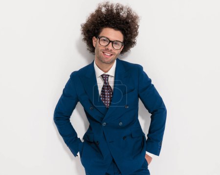 Foto de Atractivo joven hombre de negocios en elegante traje azul marino cogido de la mano en los bolsillos, siendo feliz y sonriente delante de fondo gris - Imagen libre de derechos