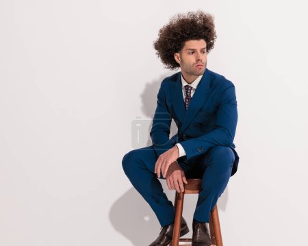 Foto de Imagen del joven reflexivo en traje azul marino sosteniendo los codos en las rodillas, mirando hacia un lado y pensando mientras está sentado sobre un fondo gris - Imagen libre de derechos