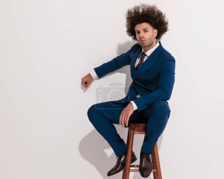 Photo for Cool elegant man with curly hair in navy blue suit sitting on wooden chair, looking forward, holding elbow on knee and punching the wall - Royalty Free Image