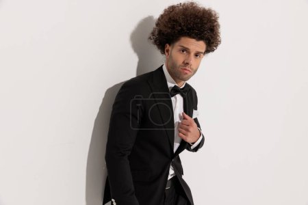 Foto de Retrato de novio elegante guapo ajustando esmoquin negro mientras mira hacia adelante en frente de fondo gris - Imagen libre de derechos
