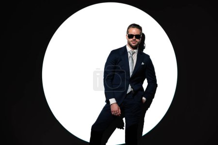 Foto de Hombre de negocios de moda confiado en traje con gafas de sol cogidas de la mano en el bolsillo y mirando hacia adelante en frente de fondo foco - Imagen libre de derechos