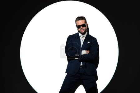 Foto de Retrato de hombre de negocios elegante y seguro de sí mismo con gafas de sol cruzando brazos y mirando hacia adelante en frente del centro de atención - Imagen libre de derechos