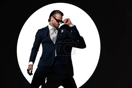 Foto de Retrato de hombre de negocios elegante sexy en traje con corbata mientras se ajustan las gafas de sol de una manera fresca frente al fondo del foco - Imagen libre de derechos