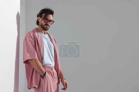 Foto de Confiado hombre casual con gafas de sol de la mano en el bolsillo, mirando a un lado y posando delante de fondo gris - Imagen libre de derechos