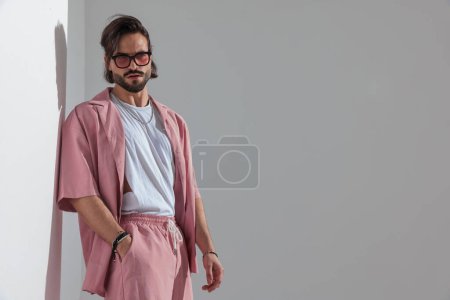 Foto de Retrato de hombre de moda casual con gafas de sol en ropa rosa cogida de la mano en el bolsillo, mirando hacia otro lado y posando frente a fondo gris - Imagen libre de derechos
