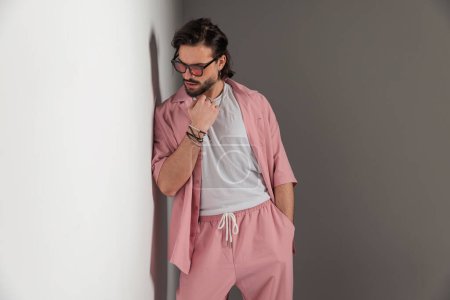 Foto de Retrato de hombre casual fresco con gafas de sol en ropa rosa cogida de la mano en el bolsillo y mirando hacia abajo mientras está acostado en una pared gris - Imagen libre de derechos