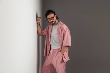 Téléchargez les photos : Portrait de jeune homme de mode avec des lunettes de soleil tenant la main dans la poche et posant tout en touchant un mur gris - en image libre de droit