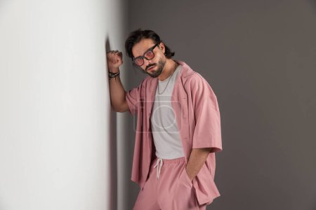 Foto de Atractivo hombre casual sin afeitar con gafas de sol de la mano en el bolsillo y tocar la pared gris en el estudio - Imagen libre de derechos
