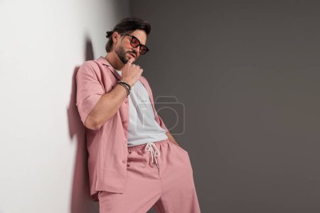 Foto de Retrato de hombre de moda sensual con gafas de sol tocando los labios y sosteniendo la mano en el bolsillo mientras está acostado en una pared en el estudio - Imagen libre de derechos