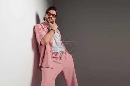 Foto de Hombre de moda casual con gafas de sol colocadas en una pared y tocando la barbilla, pensando y posando con la mano en el bolsillo sobre fondo gris - Imagen libre de derechos