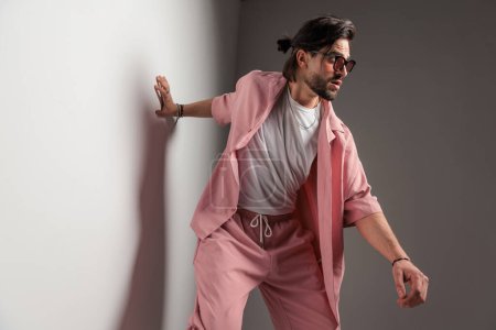 Foto de Hombre casual dinámico en ropa rosa con gafas de sol cogidas de la mano en la pared, mirando a un lado y posando de una manera fresca sobre fondo gris - Imagen libre de derechos