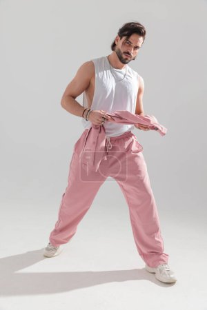 Foto de Imagen de cuerpo completo del hombre muscular sexy con ropa deportiva rosa sosteniendo hacket, mostrando los brazos y caminando delante de fondo gris - Imagen libre de derechos