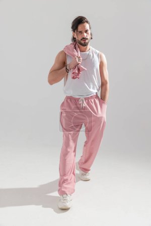 Foto de Imagen de cuerpo completo de atleta sexy chico con pantalones de color rosa de la mano en el bolsillo y la chaqueta en el hombro, mirando hacia adelante y caminando sobre fondo gris - Imagen libre de derechos
