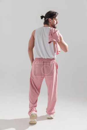 Photo for Sexy muscle man in pink gym clothes looking to side while holding jacket on shoulder and walking on grey background - Royalty Free Image