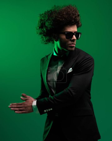 Téléchargez les photos : Homme d'affaires de mode cool en smoking noir élégant avec des lunettes de soleil et les cheveux bouclés frottant paumes sur le côté et regardant du côté opposé en face de fond vert - en image libre de droit