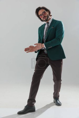 Foto de Portrait of sexy bearded man in green suit with sunglasses rubbing palms to side and posing in front of grey background - Imagen libre de derechos