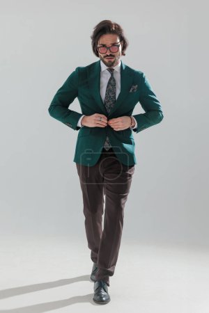 Foto de Full length picture of confident elegant man with sunglasses buttoning green suit and walking in front of grey background - Imagen libre de derechos