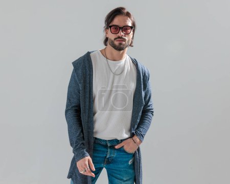 Photo for Portrait of casual young man with sunglasses wearing long hoodie holding hand in blue jeans pocket and looking forward in front of grey background - Royalty Free Image