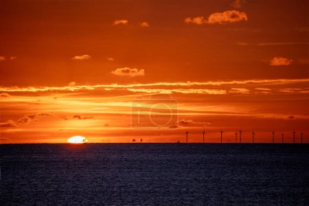 Foto de Sol sumergiéndose más allá del horizonte sobre el Canal de la Mancha con turbinas de parque eólico Rampion siluetas contra el cielo ardiente. - Imagen libre de derechos