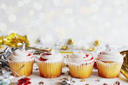 Téléchargez les photos : Quatre cupcakes à la vanille avec glaçage tourbillonnant aux fraises alignés pour une fête entre les noeuds de ruban et les confettis. Mise au point sélective avec avant-plan et arrière-plan flous. - en image libre de droit