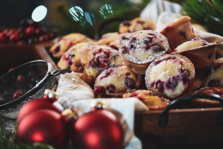 Photo for Cranberry muffins dusted with powder sugar in tray with Christmas tree and fresh berries in the background. Ornaments and vintage duster nearby. Selective focus with blurred foreground and background. - Royalty Free Image
