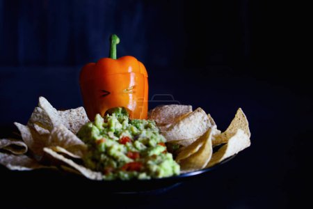 Photo for Stuffed sweet orange bell pepper as monster with cut out face carved into a Halloween pumpkin Jack O'Lantern throwing up guacamole served with chips. Selective focus on mouth with blurred foreground and background. - Royalty Free Image