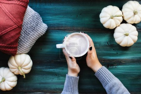 Photo for Flatlay of woman's hands holding hot, steaming cup of coffee over a rustic green table with autumn sweaters and white pumpkins. Table top view. Overhead. - Royalty Free Image