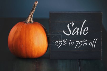 Photo for Autumn clearance for sale black wooden sign with 25 to 75% off. Orange pumpkin on a green table. Selective focus with blurred foreground and background. - Royalty Free Image