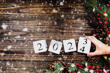 Photo for Woman's hand flipping New Year's 2021 wood calendar blocks to 2023. Christmas tree lights, pine branches, red winter berries and snow. Top view. - Royalty Free Image