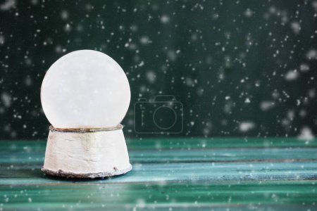 Photo for Magical empty snowglobe over a green rustic table background with falling snow. Blurred foreground and background. Copy space available. - Royalty Free Image