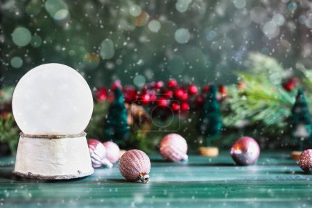 Photo for Magical empty snowglobe over a green rustic table background with falling snow. Christmas decorations in the background. Blurred foreground and background. Copy space available. - Royalty Free Image