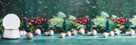 Photo for Banner of a magical empty snowglobe over a green rustic table background with falling snow. Christmas decorations in the background. Blurred foreground and background. Copy space available. - Royalty Free Image