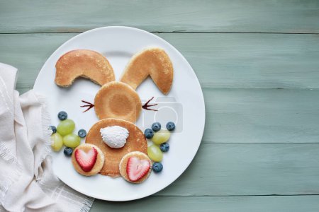 Photo for Fun, creative pancake bunny rabbit with fluffy tail hopping away for children with whipped cream and strawberries with green grapes and blueberries as Easter eggs to encourage kids to eat healthy food - Royalty Free Image