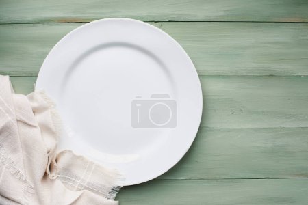 Photo for An empty white plate with napkin over a rustic green spring table, shot from flat lay or table top view position. Free space for text. - Royalty Free Image