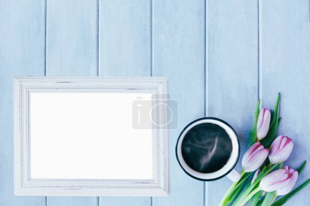 Photo for Blank empty picture frame with hot coffee and tulip flowers over blue rustic background with spring flowers. Table top view. - Royalty Free Image
