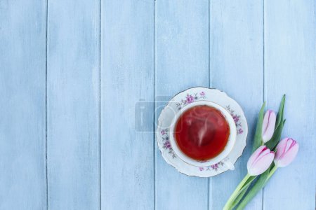Photo for Steaming hot cup of tea and pink spring tulip flowers over a rustic blue wooden table. Table top view. Directly above. - Royalty Free Image
