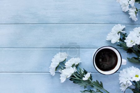 Photo for White enamel cup of hot steaming coffee surrounded by white Chrysanthemum flowers over a blue rustic table. Table top view. Directly above. - Royalty Free Image