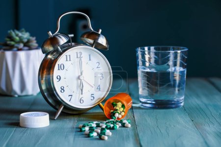Photo for Open bottle of  prescription pills by an alarm clock and a glass of water on wooden night stand table. Selective focus with blurred background. - Royalty Free Image