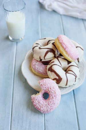 Photo for Frosted vanilla donuts with chocolate swirls and strawberry pink doughnuts with coconut flakes. Bite missing. Glass of milk in background. Selective focus with blurred foreground and background. - Royalty Free Image