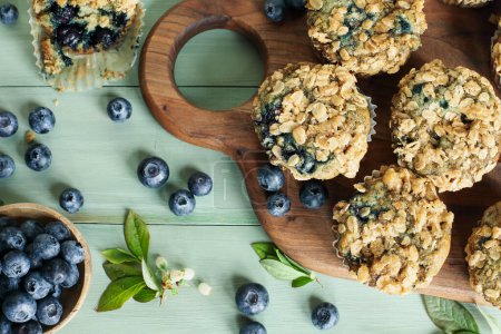 Photo for Fresh blueberry muffins with oat streusel topping and raw blueberries spilling from a wooden spoon. Table top view with focus on muffin tops and blurred background. - Royalty Free Image