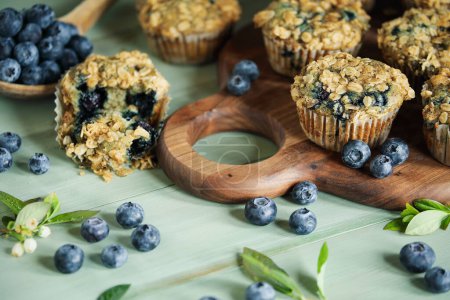 Photo for Fresh blueberry muffins with oat streusel topping and raw blueberries spilling from a wooden spoon. Selective focus on muffins on cutting board with blurred background. - Royalty Free Image